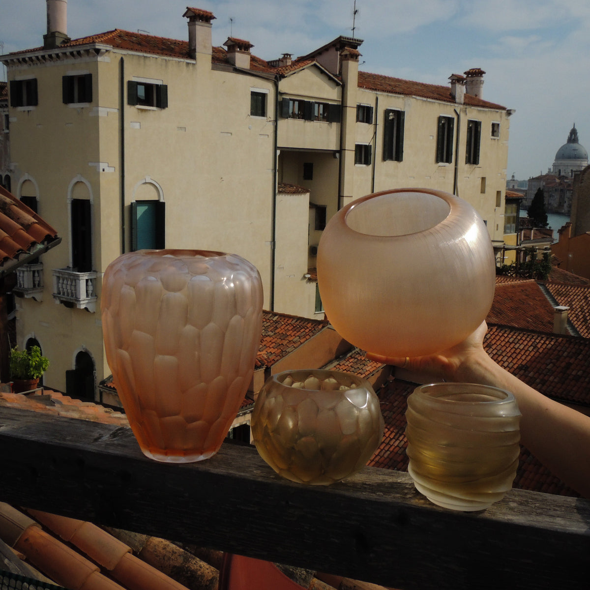 Riccio Rosé Millerighe vase held in hand, shown with other cold-carved Murano glass vases on a wooden ledge in Venice